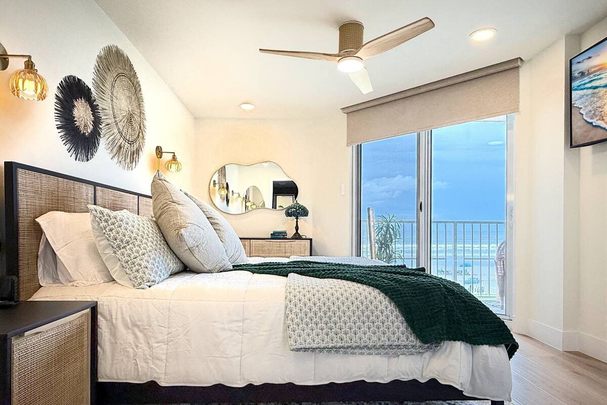 Une chambre avec un grand lit, un ventilateur de plafond, une vue sur l’océan par une grande fenêtre et un balcon.