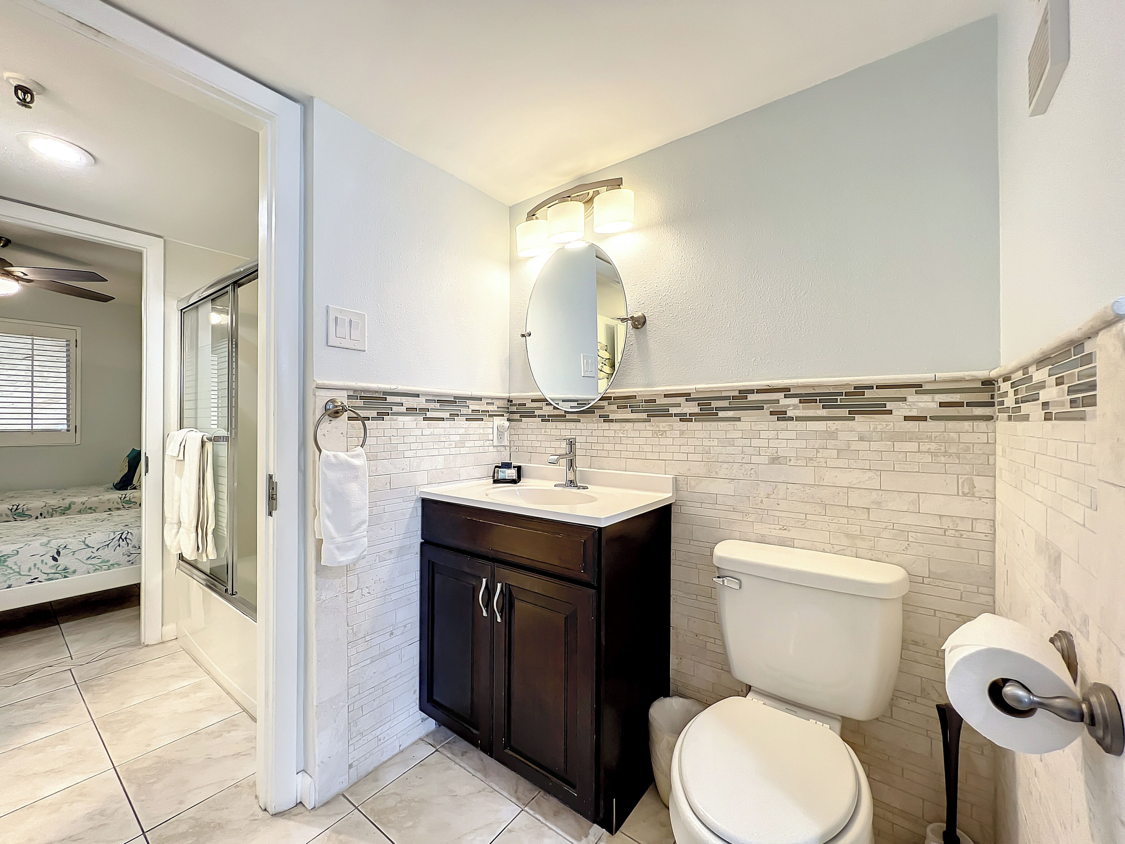 A bathroom with a toilet, sink, and mirror. There is a towel rack with a towel hanging on it. The bathroom has a tiled floor and a window with blinds.
