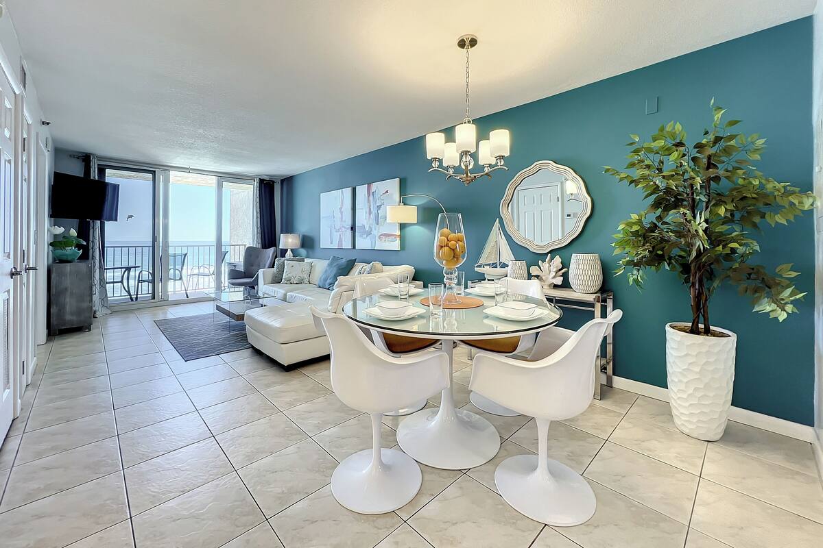 A modern dining area with a glass table, white chairs, and a teal accent wall.
