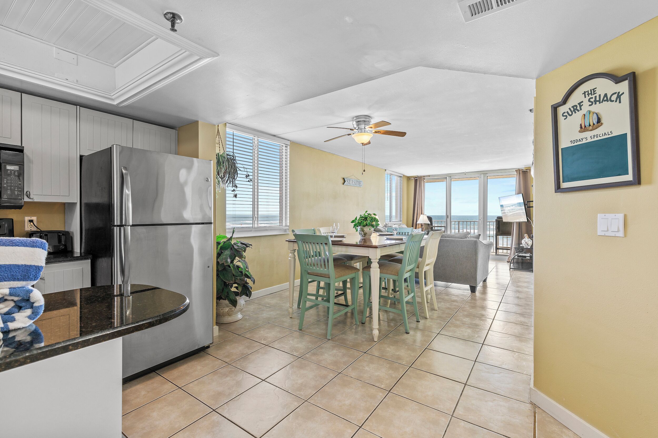 A modern kitchen with a stainless steel refrigerator, a dining area with green chairs, and a ceiling fan.