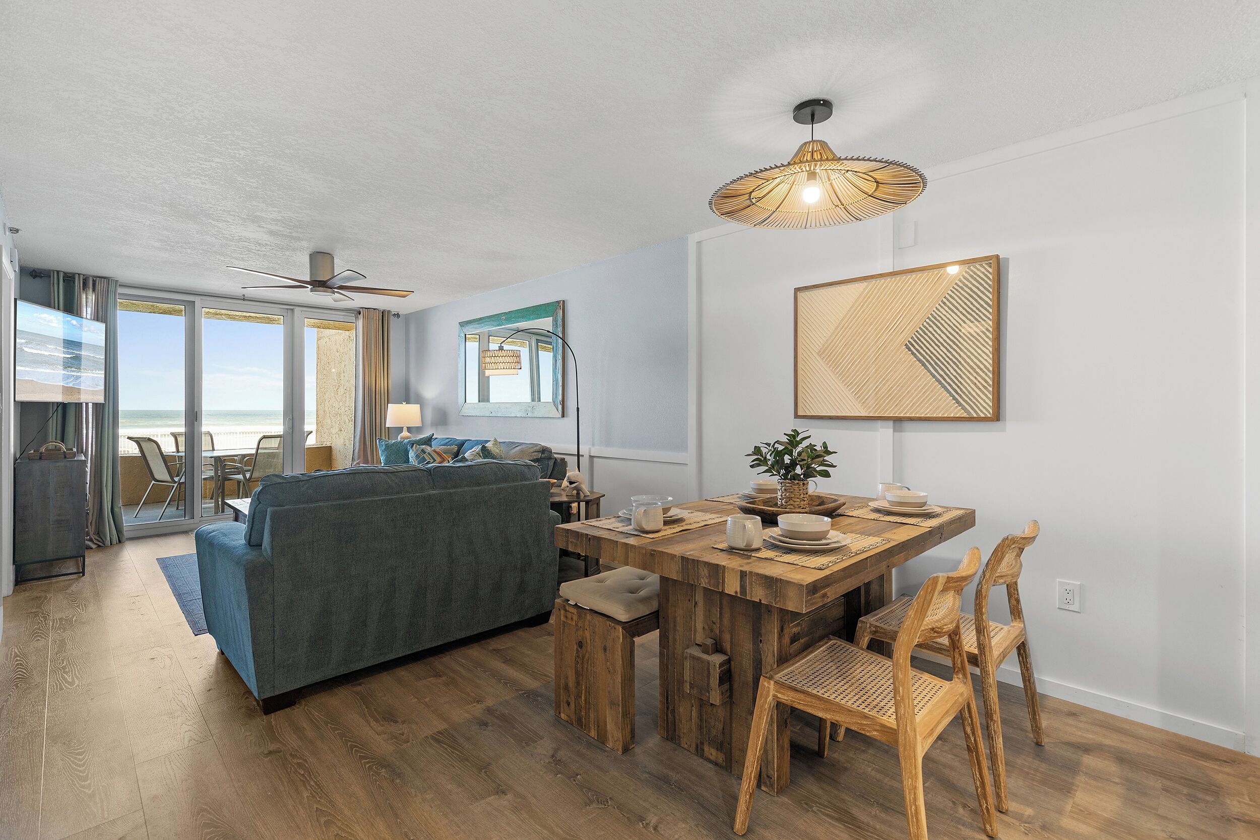 Una sala de estar moderna con sofá, mesa de comedor y sillas. Se observa un ventilador de techo, un ventanal con vistas y una lámpara fijada a la pared.