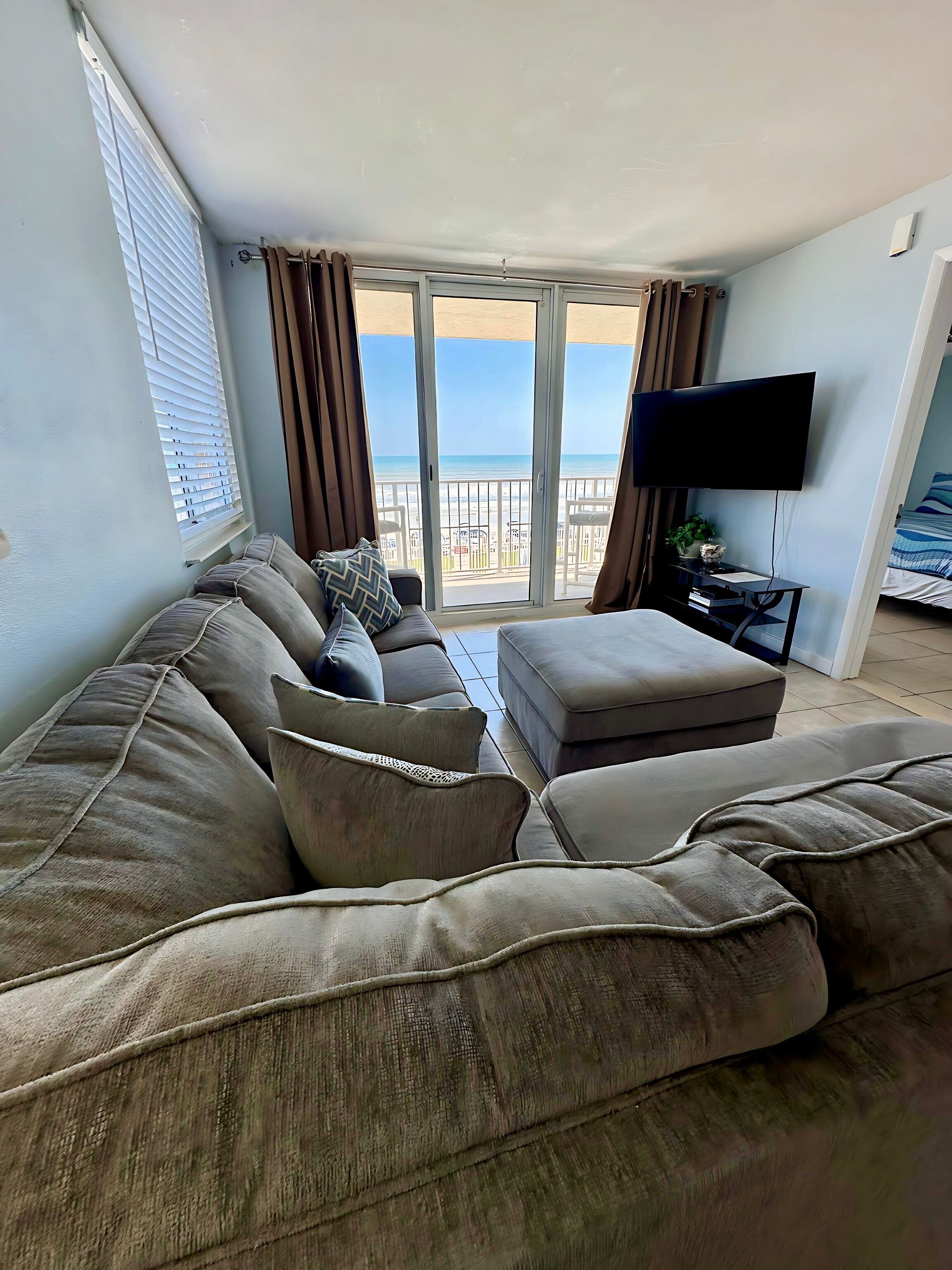Un soggiorno con un divano ampio, una televisione e un balcone con vista sulla spiaggia.