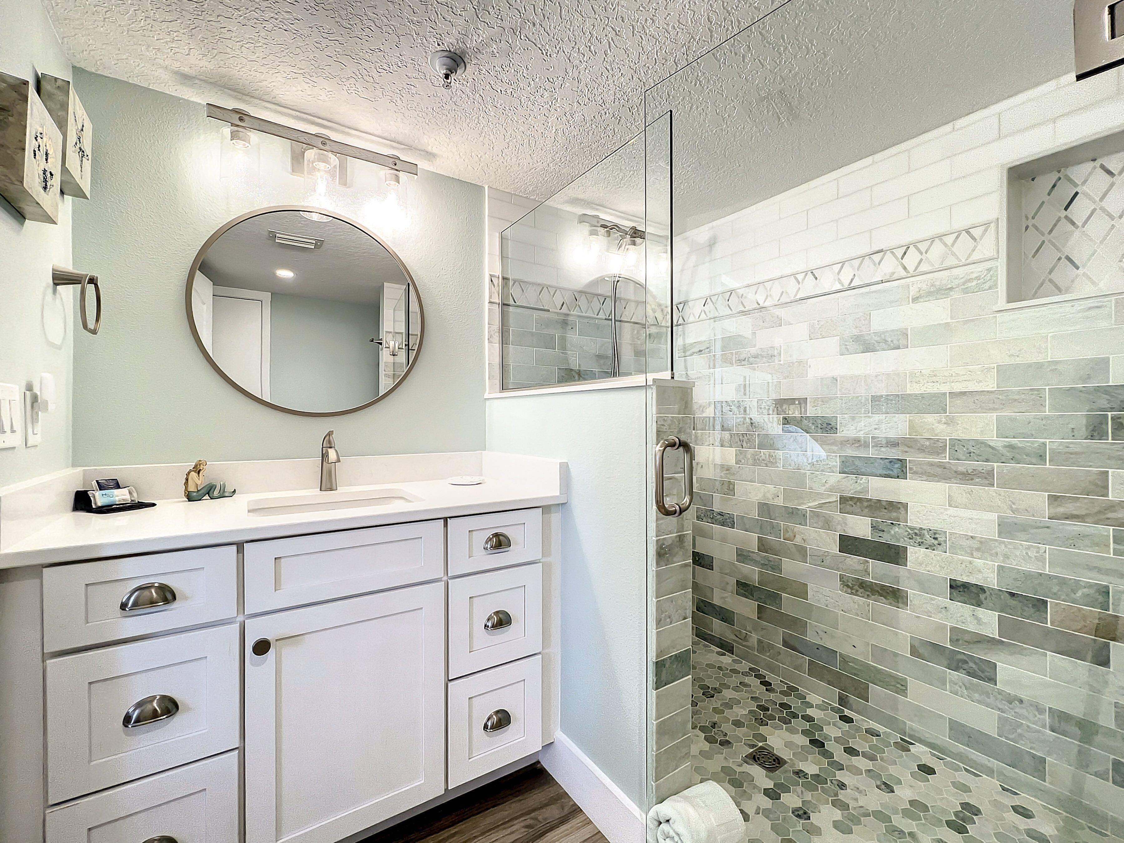 A bathroom with a white vanity, a round mirror, and a shower with mosaic tiles.