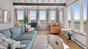 Un salón amplio con un sofá azul, un sillón naranja y una mesita. Amplias ventanas ofrecen vistas al océano.