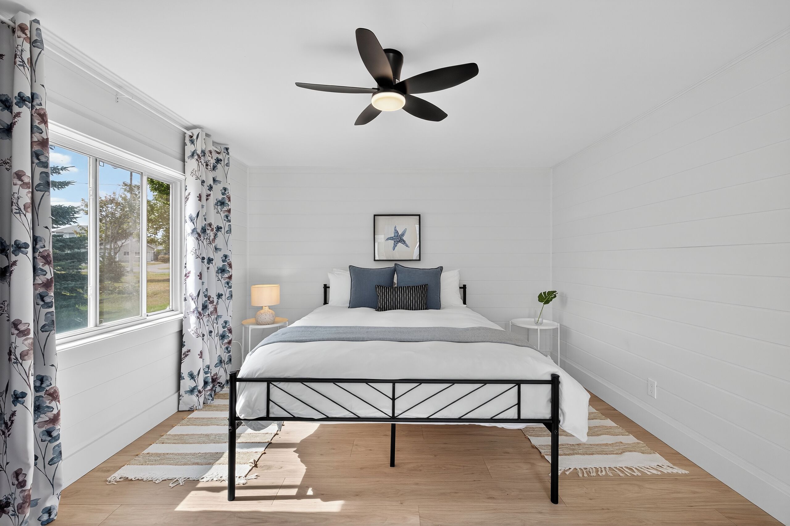 Une chambre à coucher avec un lit, un ventilateur de plafond, une fenêtre avec des rideaux, une table de chevet et un tableau encadré au mur.
