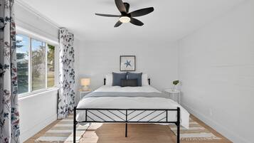 Une chambre à coucher avec un lit, un ventilateur de plafond, une fenêtre avec des rideaux, une table de chevet et un tableau encadré au mur.