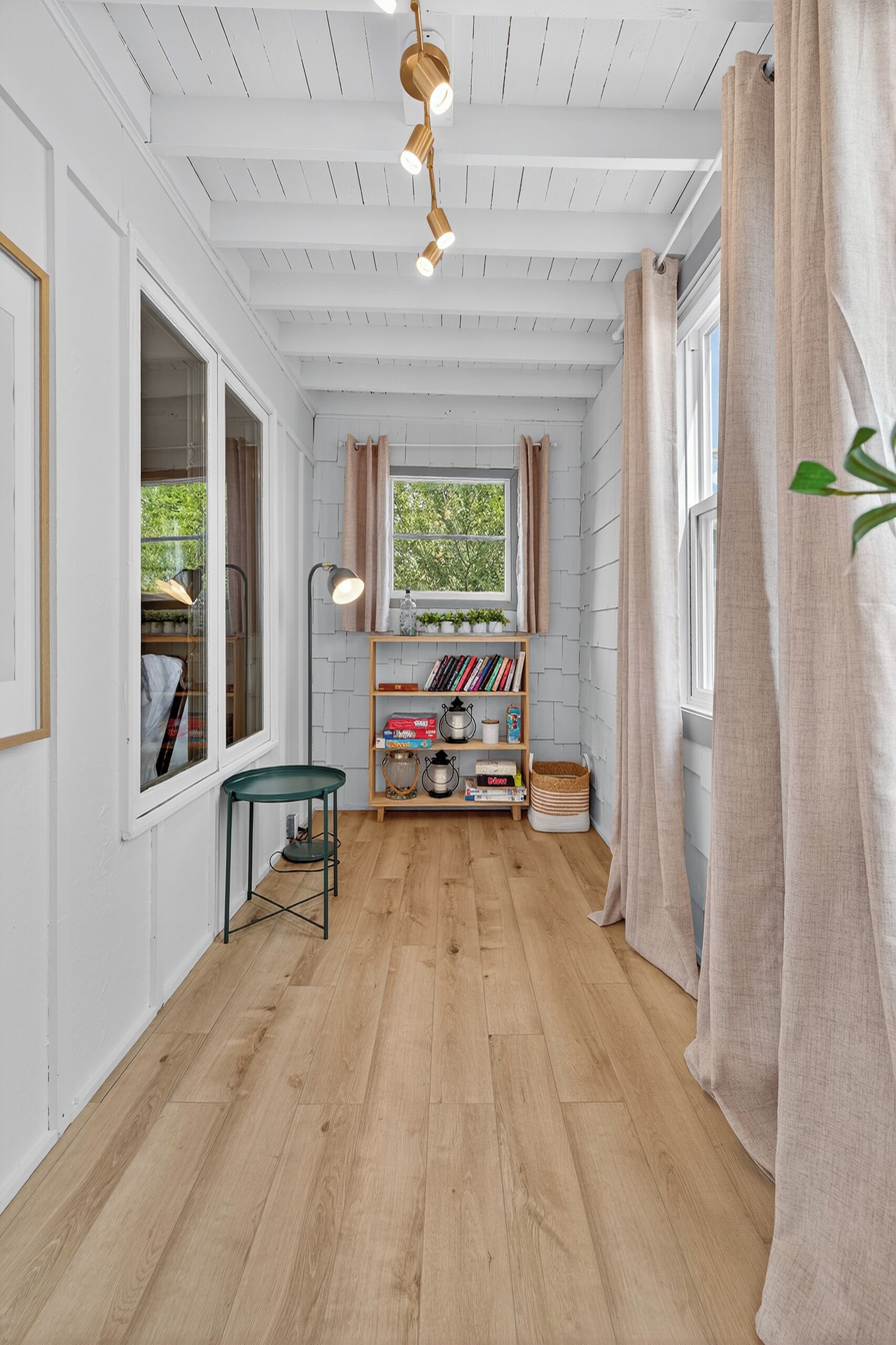 Un couloir avec un plancher en bois, une bibliothèque, une table et une fenêtre avec des rideaux.