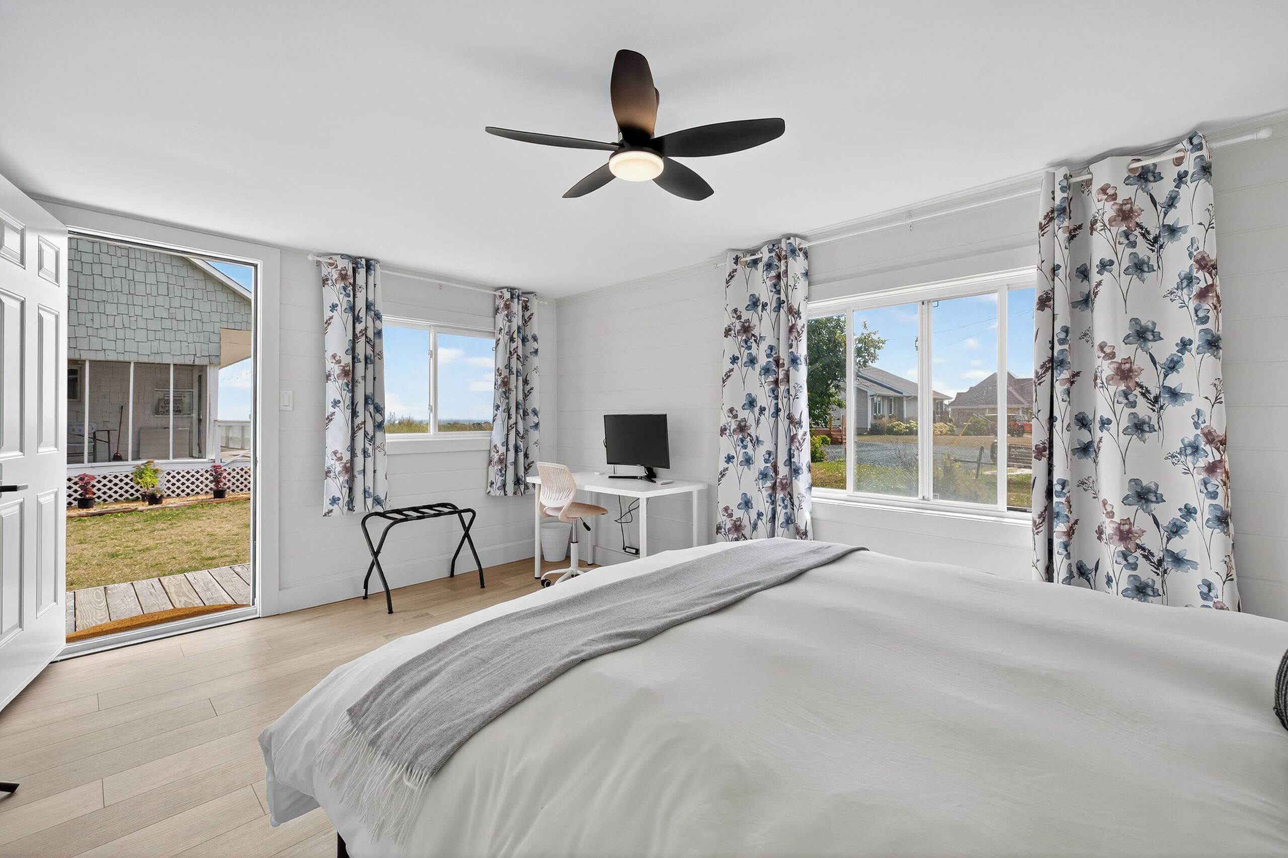 A bedroom with a bed, a desk, a chair, a ceiling fan, and large windows with curtains.