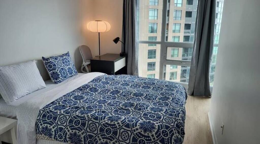 A hotel room with a large bed, a desk, and a chair. There is a window with a view of buildings.