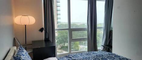 A bedroom with a bed, a nightstand, a lamp, and a window with a view of greenery and buildings.