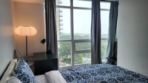 A bedroom with a bed, a nightstand, a lamp, and a window with a view of greenery and buildings.