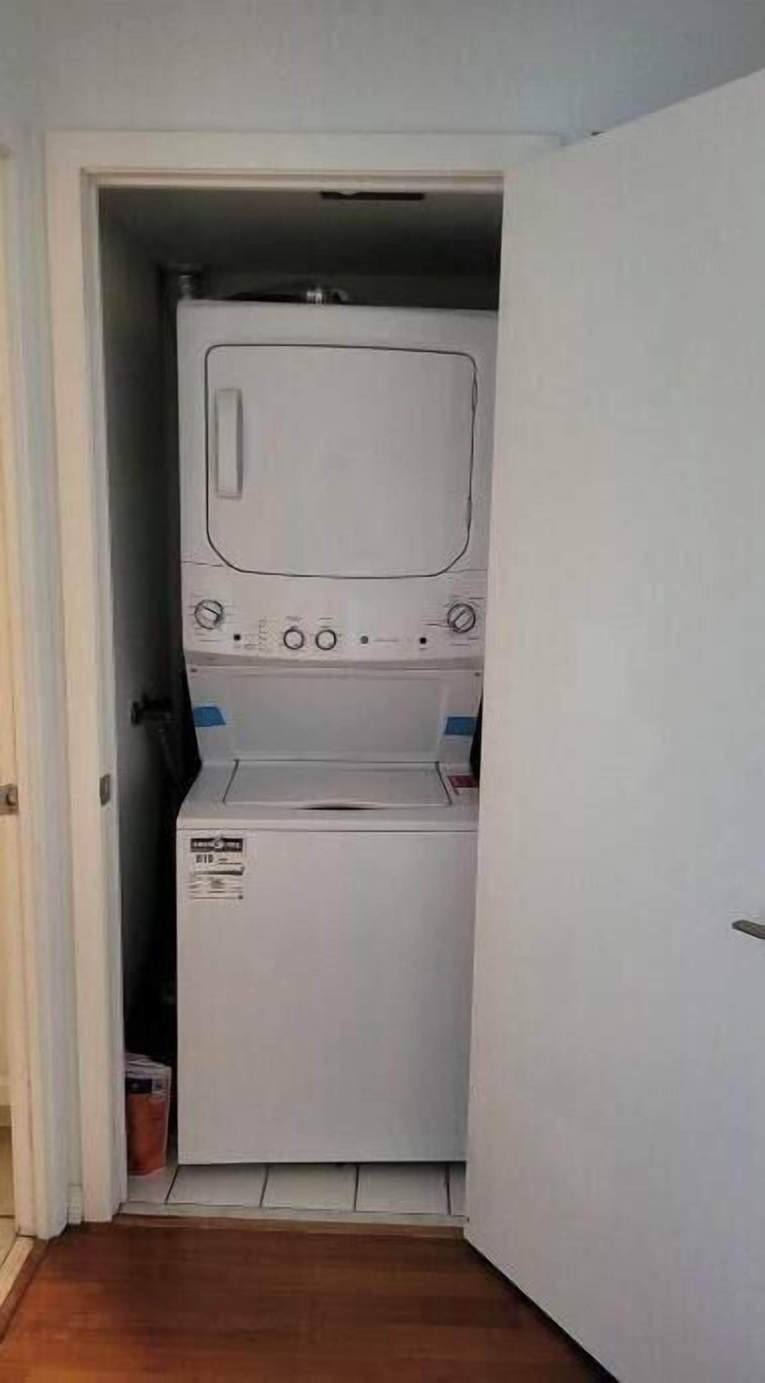 A compact laundry room with a washing machine and dryer, a small trash bin, and a door leading to another room.