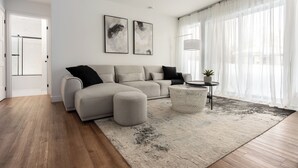 Un salón moderno con un sofá seccional gris, una mesa redonda de centro y un ventanal con cortinas translúcidas.