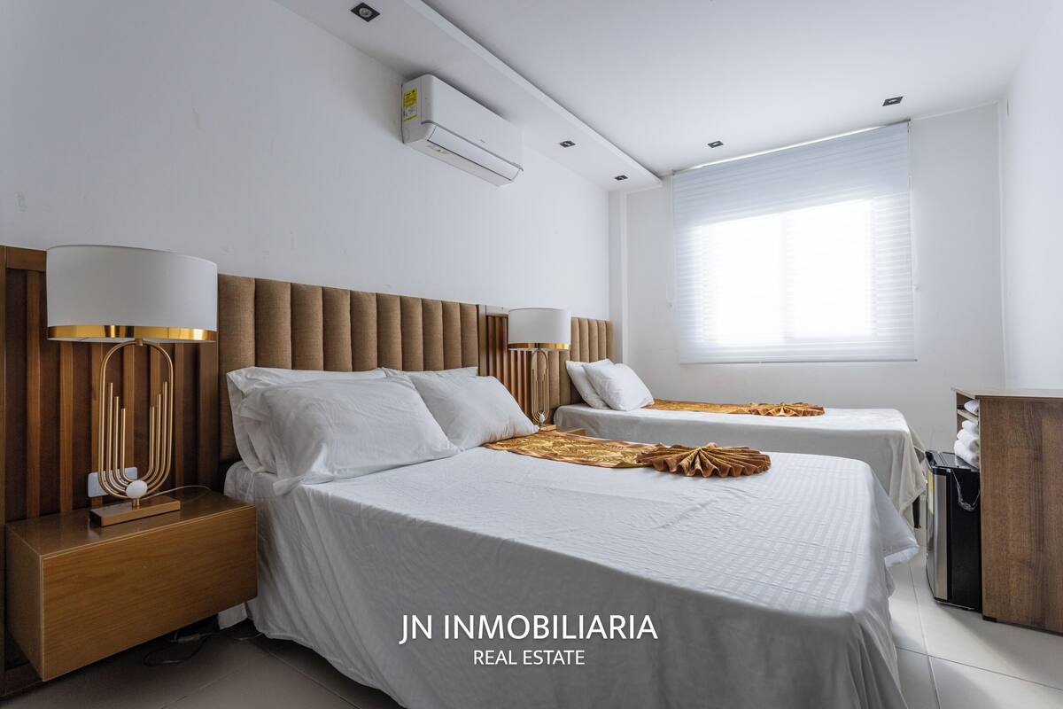 Een moderne hotelkamer met twee bedden, een houten hoofdbord, bedlampen, een airconditioningsysteem en een raam met jaloezieën.