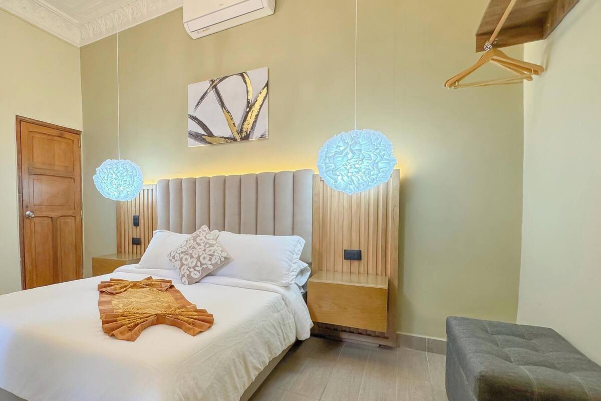 Een netjes opgemaakt bed met een witte deken en een opgevouwen bruine plaid, een houten hoofdbord, en twee wandlampjes met decoratieve verlichting.