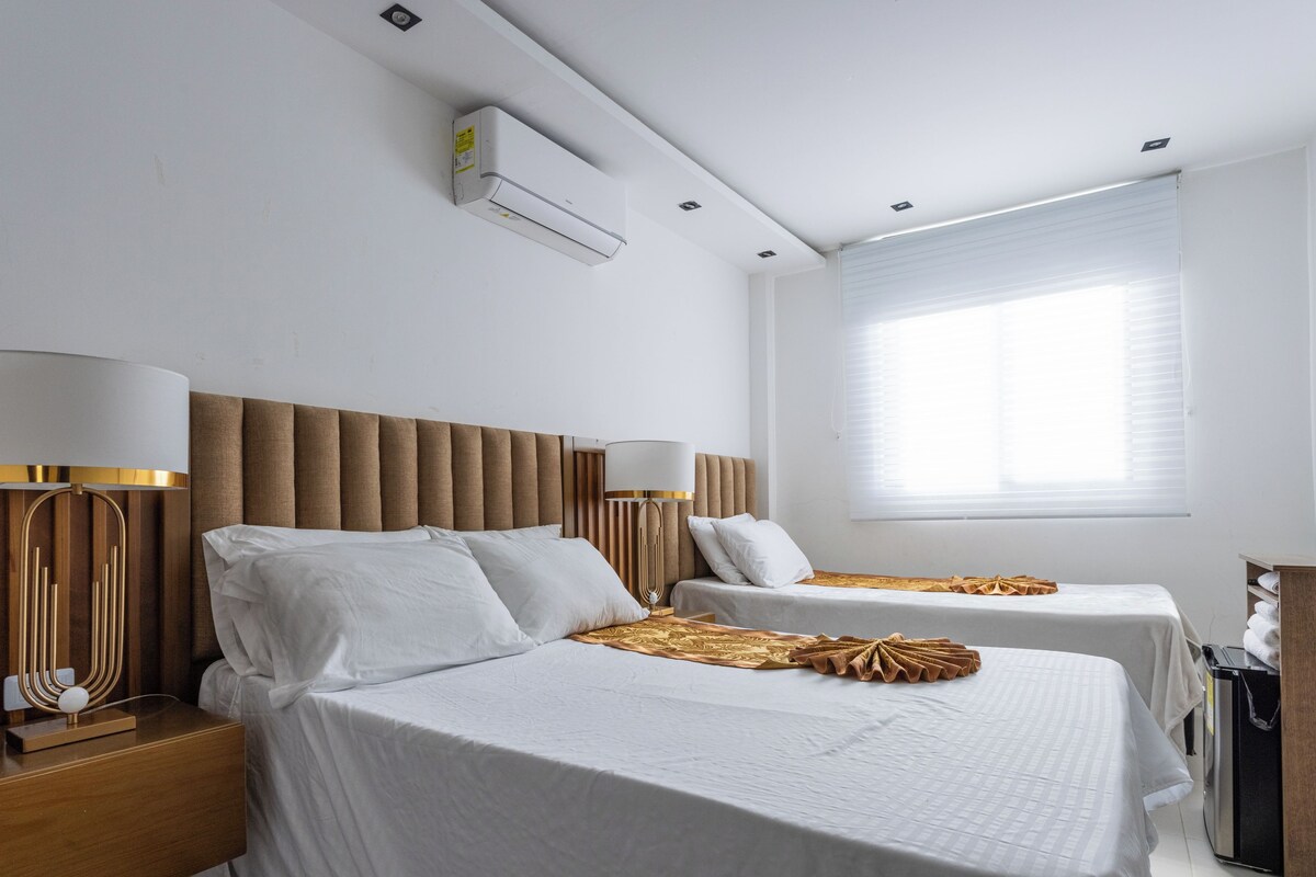 A modern hotel room with two beds, a wooden headboard, bedside lamps, an air conditioning unit, and a window with blinds.