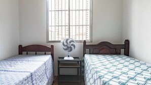 A small room with two single beds, a standing fan, and a window with barred glass. - Apto Pet-friendly a 400m da Praia do Boqueir o (Praia Grande)