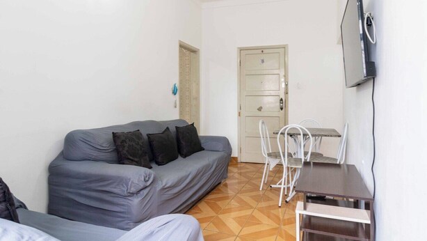 A small, compact room with a grey sofa, a small table with chairs, a TV mounted on the wall, and a door leading to another room. - Apto Pet-friendly a 400m da Praia do Boqueir o (Praia Grande)