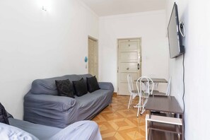 A small, compact room with a grey sofa, a small table with chairs, a TV mounted on the wall, and a door leading to another room. - Apto Pet-friendly a 400m da Praia do Boqueir o (Praia Grande)