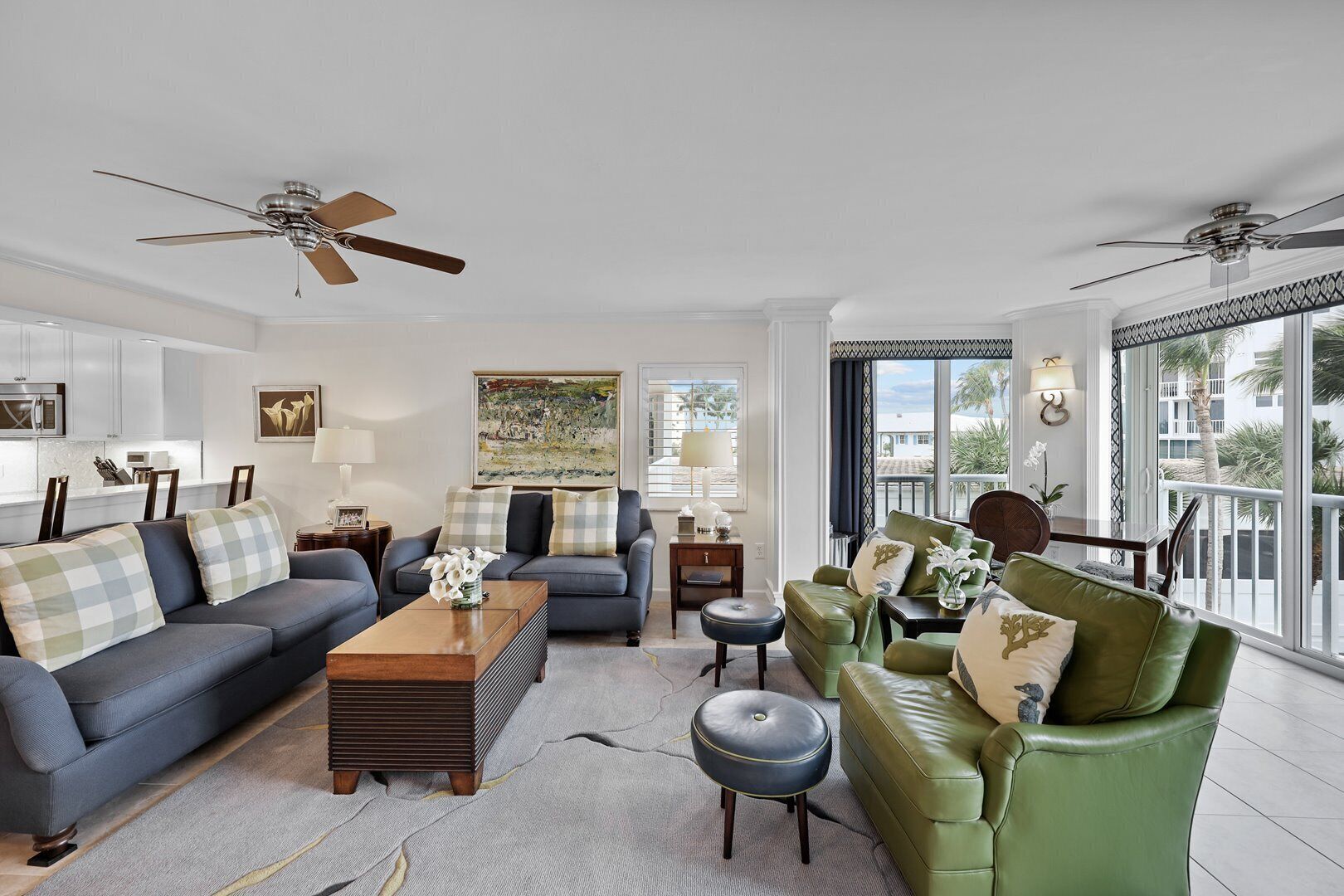 A spacious living room with two sofas, a coffee table, and a green armchair. There are ceiling fans, a large window with a view, and a painting on the wall.