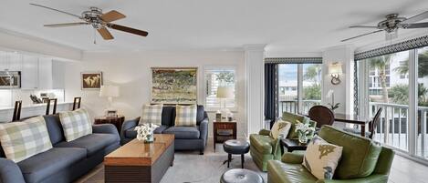 A spacious living room with two sofas, a coffee table, and a green armchair. There are ceiling fans, a large window with a view, and a painting on the wall.