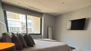 Habitació d'hotel moderna amb un llit gran, una taula de nit amb una làmpada, una televisió de pantalla plana penjada a la paret i vistes a l'exterior a través d'una finestra gran.