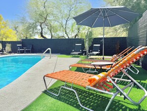 Piscina com espreguiçadeiras e mesa próximas, cercada por um muro de tijolos e vegetação.
