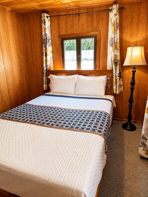 A wooden-paneled room with a bed, curtains, a window, and a lamp.
