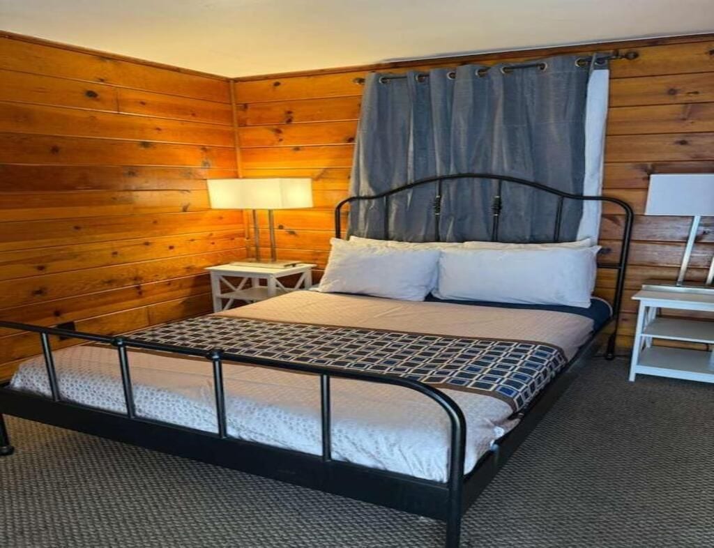 Una cama individual con estructura metálica, una mesita de noche, una lámpara y una cortina.