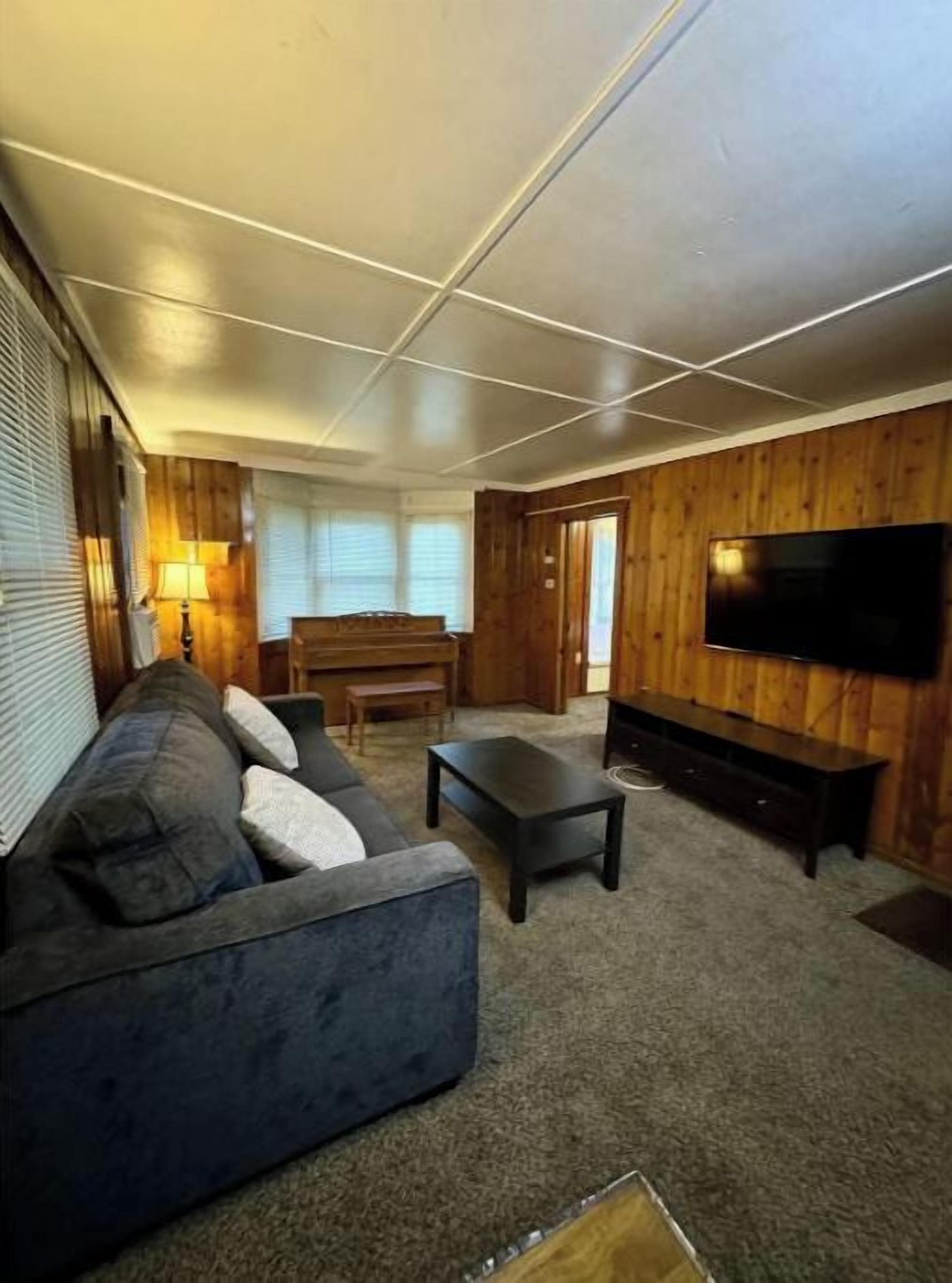 A living room with a sectional sofa, a coffee table, a TV mounted on a wooden wall, and a window with blinds.