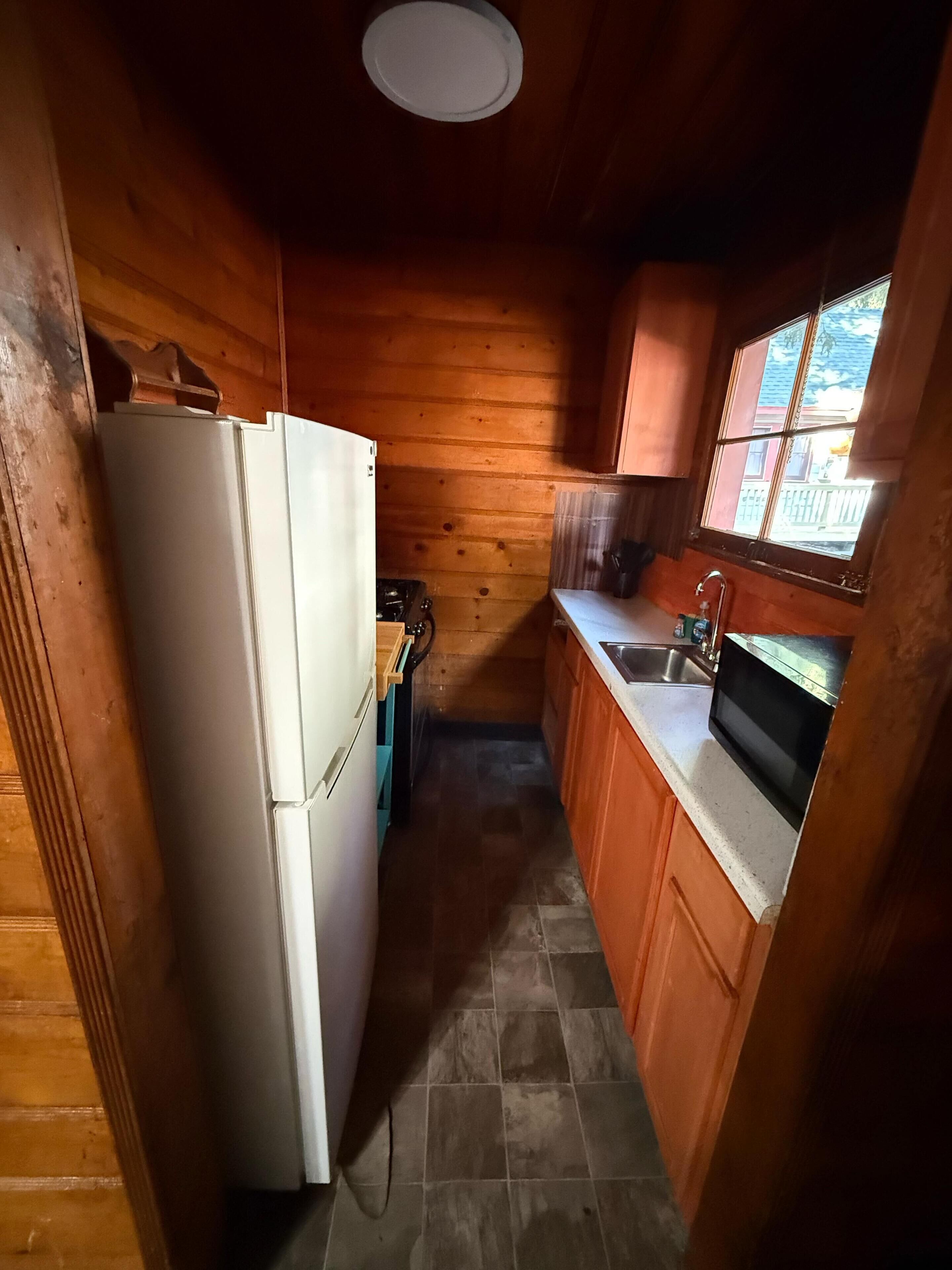A compact kitchen with wooden walls, a refrigerator, a stove, a sink, and a window.