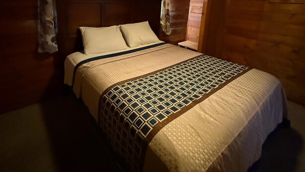A dimly lit room with a wooden ceiling, a bed with a patterned bedspread, and floral curtains. - Crest Lodge Historic Mountain Inn - ArrowBear (Crestline)