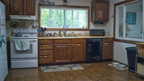 A kitchen with wooden cabinets, a stove, a sink, and a window with a view of trees. - Loon Lodge on Bear Lake (Bear Lake)