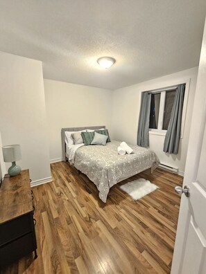 Um quarto com cama, mesa de cabeceira, luminária, janela com cortinas e porta.