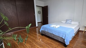 A neatly made bed with a blue blanket and white pillows, a small round table with a plant, and a potted plant with green leaves.