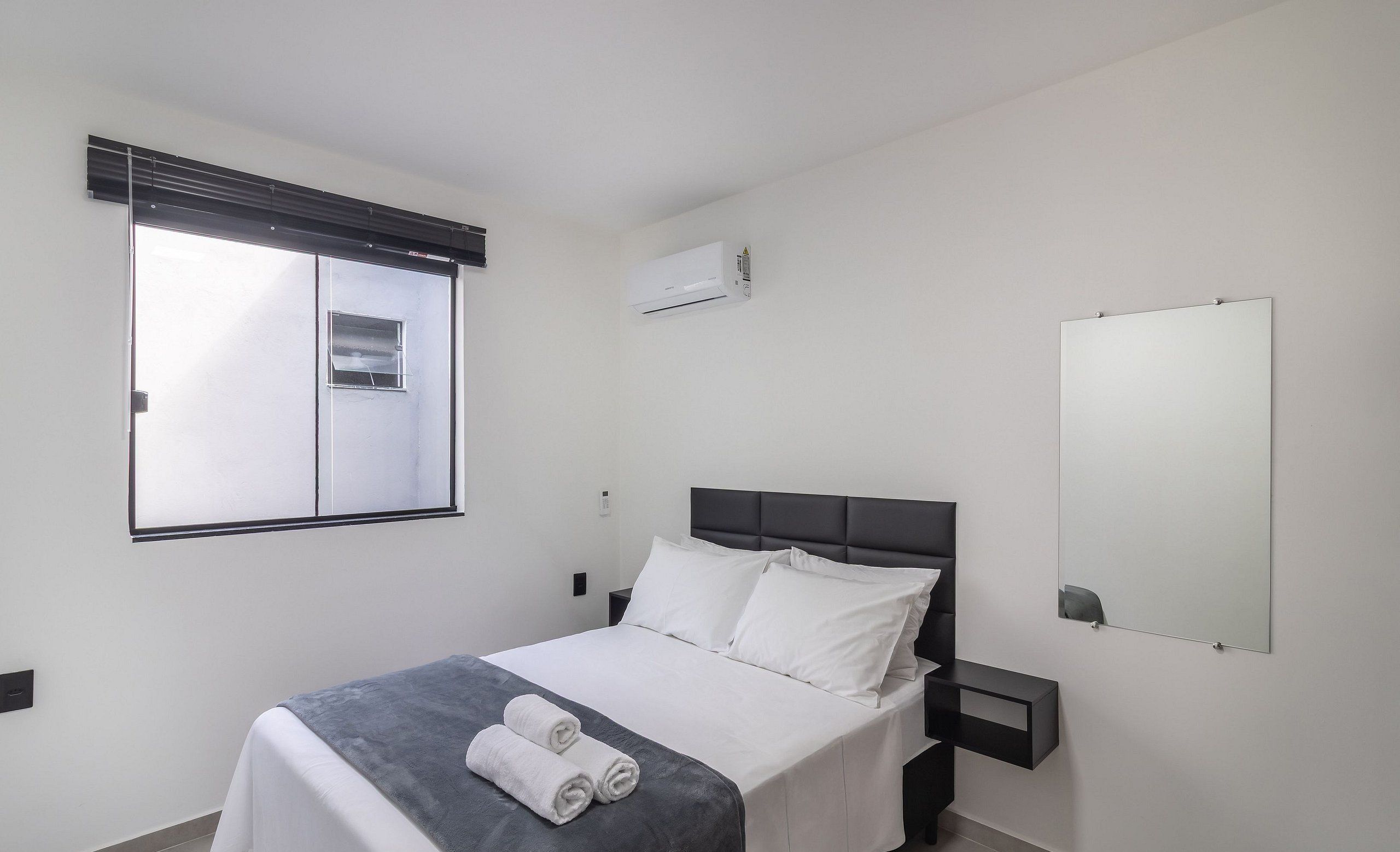 Une chambre à coucher moderne comprenant un lit, une table de chevet, une commode, une fenêtre avec des stores, un climatiseur et un miroir mural.