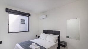 A modern bedroom with a bed, a bedside table, a nightstand, a window with blinds, an air conditioning unit, and a mirror on the wall. - Complete and Well-located Studio, Blumenau Lfh0306 (Blumenau)