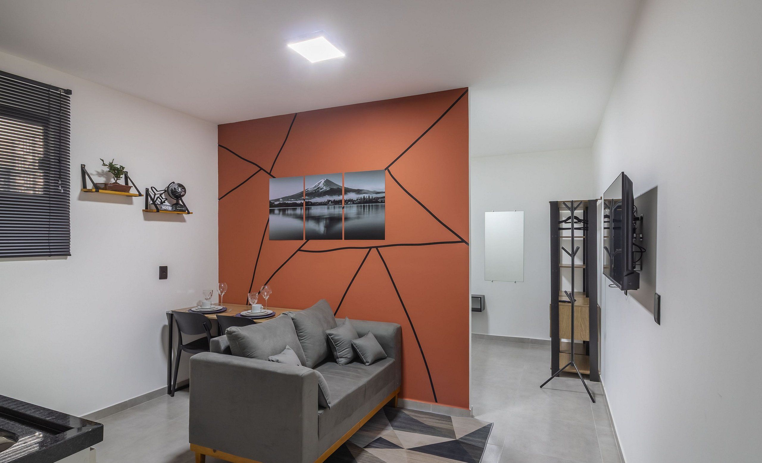 Un salon moderne avec un canapé gris, une table à manger avec des chaises, un téléviseur posé sur un meuble et un motif géométrique sur le mur.