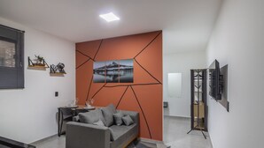 A modern living room with a grey sofa, a dining table with chairs, a TV on a stand, and a geometric wall design. - Complete and Well-located Studio, Blumenau Lfh0306 (Blumenau)