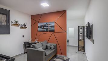 A modern living room with a grey sofa, a dining table with chairs, a TV on a stand, and a geometric wall design.