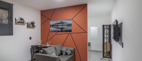 Ein modernes Wohnzimmer mit einem grauen Sofa, einem Esstisch mit Stühlen, einem Fernseher auf einem Lowboard und einem geometrischen Wanddesign.
