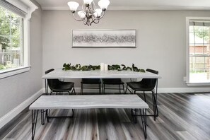 A dining area with a white table, black chairs, a bench, a chandelier, and a framed abstract painting on the wall.