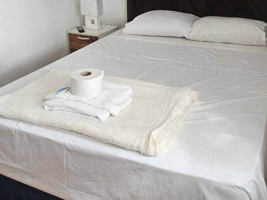 A neatly made bed with white linens, a rolled-up towel, and a remote control on a nightstand beside it.