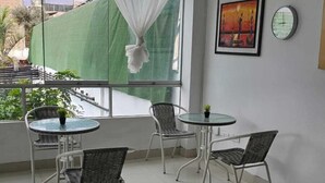 A balcony with a glass table and chairs, a white curtain, and a view of buildings and greenery. - PACHA RODEO (Pachacamac)