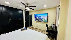 A hotel room with a bed, a desk with a computer, a TV on the wall, and a ceiling fan. - Brooklyn Loft  (Sosúa)