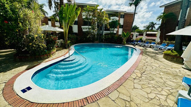 A swimming pool with a brick border, surrounded by lounge chairs and umbrellas, with a building in the background. - Brooklyn Loft  (Sosúa)
