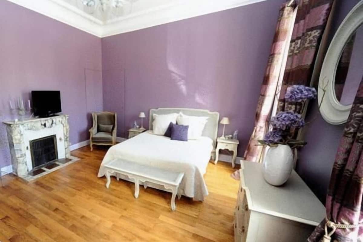 A bedroom with a bed, a nightstand, a chair, a fireplace, and a mirror.