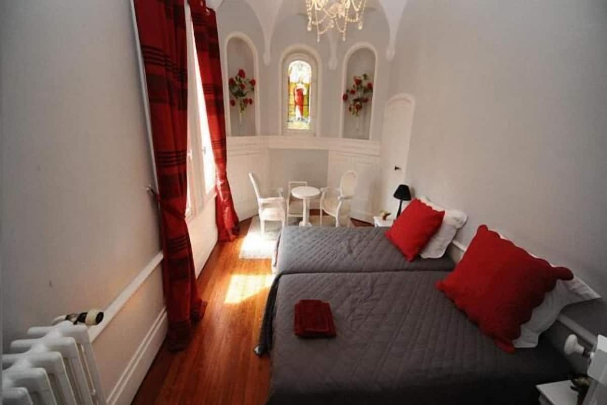 A bedroom with a bed, a chair, a table, and a window with flowers.