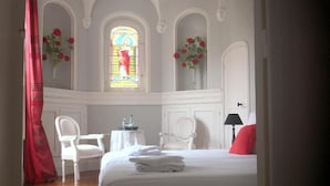 A hotel room with a bed, two chairs, a small table, a vase with flowers, and a stained glass window. - la  demeure de Laclais (Le Mans)