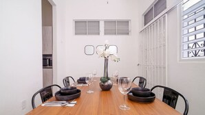 Une table à manger avec des bols noirs, des verres à vin et un vase rempli de fleurs. Il y a deux fenêtres avec des cadres blancs et une porte blanche avec un portail en métal.